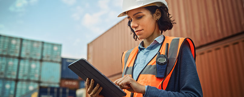 Smiling Portrait of a Beautiful Latin Female Industrial Engineer in White Hard Hat, High-Visibility Vest Working on Tablet Computer. Inspector or Safety Supervisor in Container Terminal.