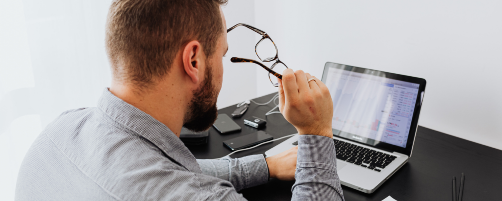 A man sits at a desk while using a laptop computer, facing a grey wall